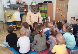 Widok na pszczelarza w kapeluszu z siateczką oraz słuchające jego opowieści przedszkolaki.